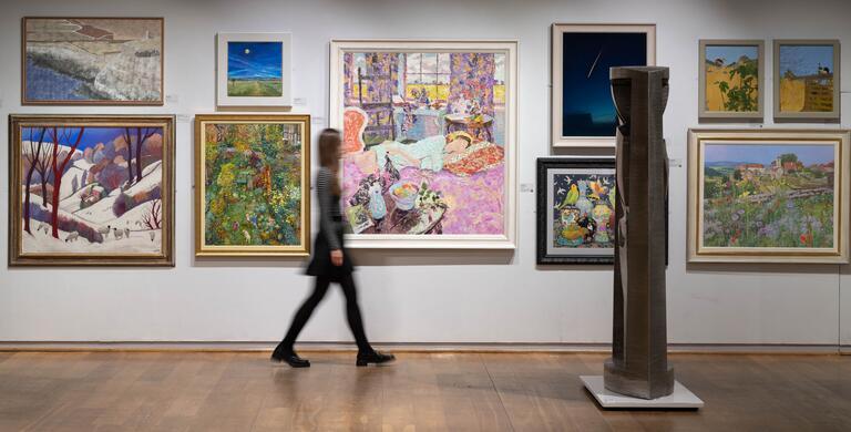 A visitor walks through the gallery, passing by a salon hang of colourful wall-mounted artworks and towards a large, floor-based sculpture.
