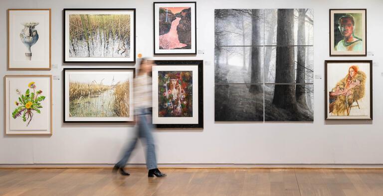A visitor walks through the gallery space, passing a gallery wall featuring water colour paintings of varied subjects.