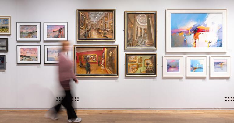 A visitor walks through the gallery space, passing a gallery wall featuring oil paintings of varied subjects.