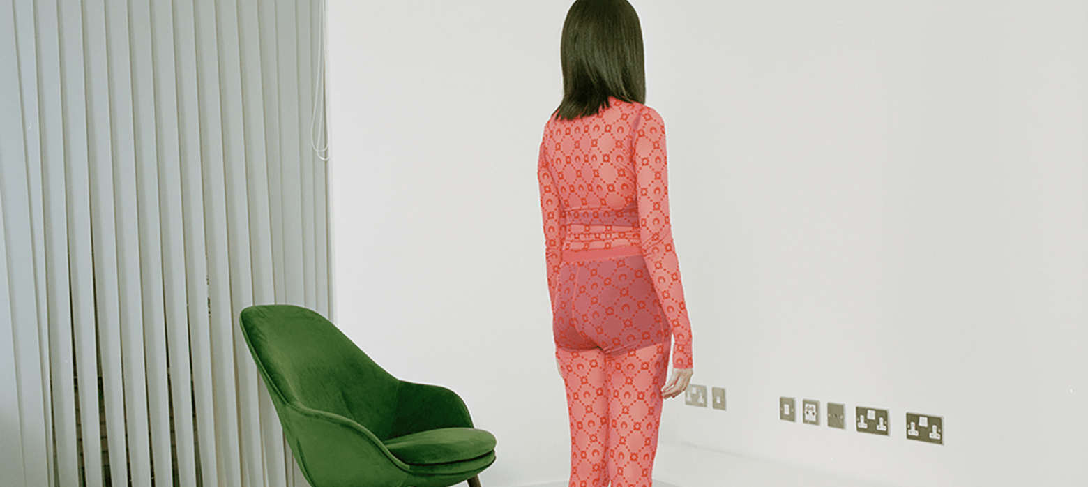 photo of a woman in a red mesh jumpsuit facing a wall with a green chair next to her