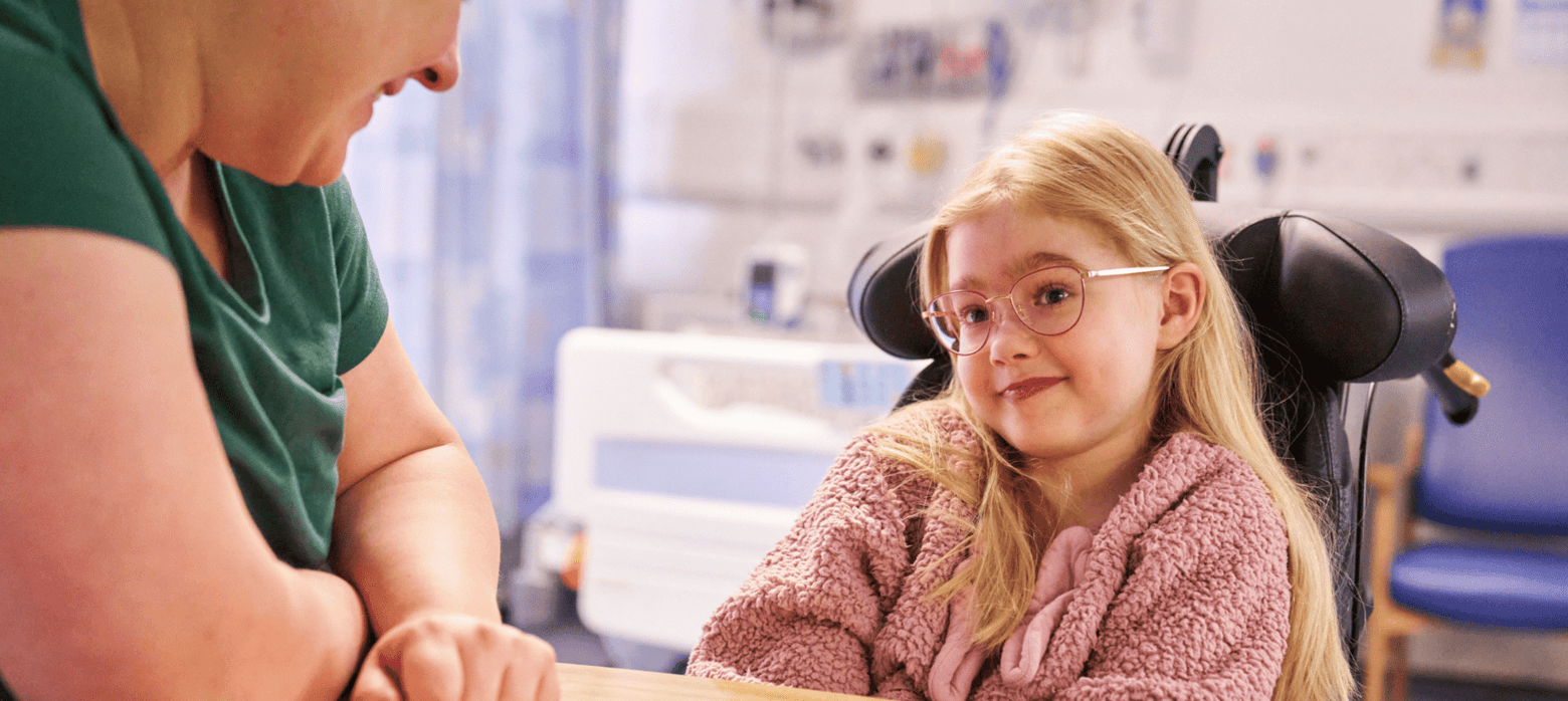 a woman and a child with blonde hair, oink cardigan and pink glasses sat in a hospital room
