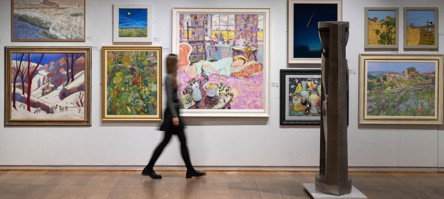 A visitor walks through the gallery, passing by a salon hang of colourful wall-mounted artworks and towards a large, floor-based sculpture.