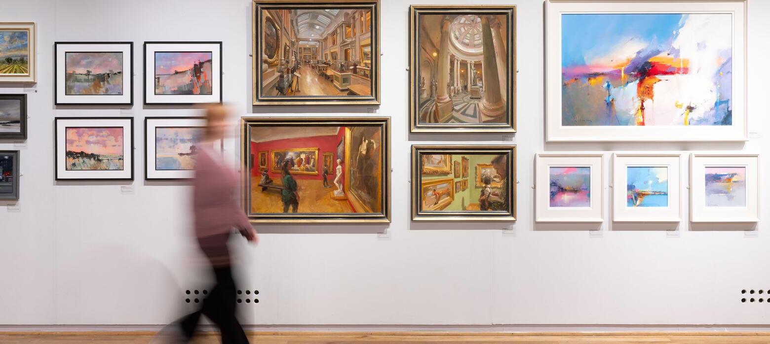 A visitor walks through the gallery space, passing a gallery wall featuring oil paintings of varied subjects.