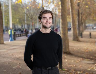 A white man with brunette hair and black top stands in front of trees on The Mall