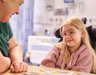 a woman and a child with blonde hair, oink cardigan and pink glasses sat in a hospital room