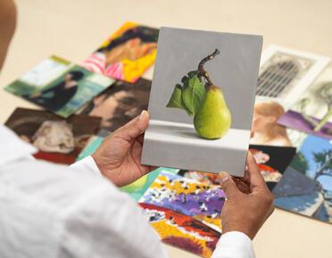 Person holding postcard sized oil painting of a pear with other artworks in the background