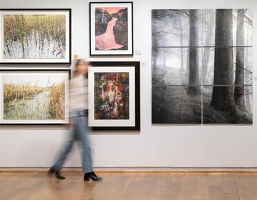 A visitor walks through the gallery space, passing a gallery wall featuring water colour paintings of varied subjects.