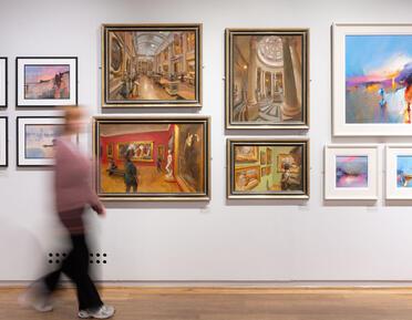 A visitor walks through the gallery space, passing a gallery wall featuring oil paintings of varied subjects.